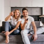 Un couple surpris et souriant regarde une émission passionnante via leur service iptv, assis sur un canapé gris. L'homme tient la télécommande.