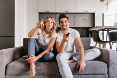 Un couple surpris et souriant regarde une émission passionnante via leur service iptv, assis sur un canapé gris. L'homme tient la télécommande.
